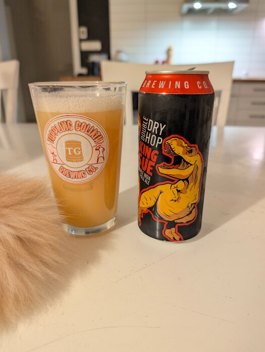 Ölglas med Toppling Goliath logga och en King Sue burk på ett bord, delvis skymd av en kattsvans.