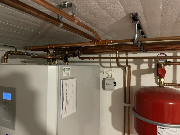 Rörsystem ovanför en vit värmepump med flera kopparrör, en röd expansionstank och en tryckmätare i ett teknikrum.