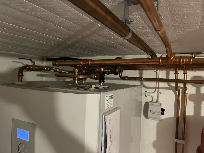 Rörsystem över en värmepump med flera ventiler och kopparrör, fokus på svart vred nära taket för påfyllning av vatten.