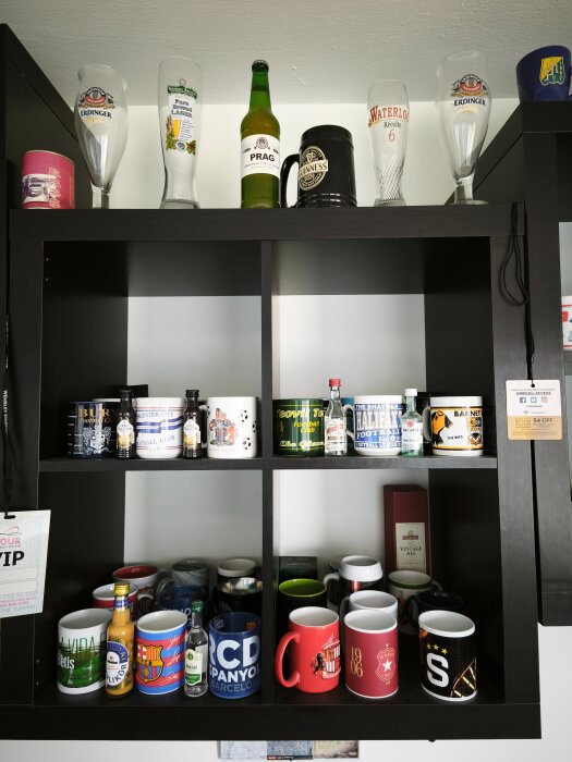 Samling av fotbollsmuggar och glas på en hylla, inklusive olika klubblag och ölflaskor.