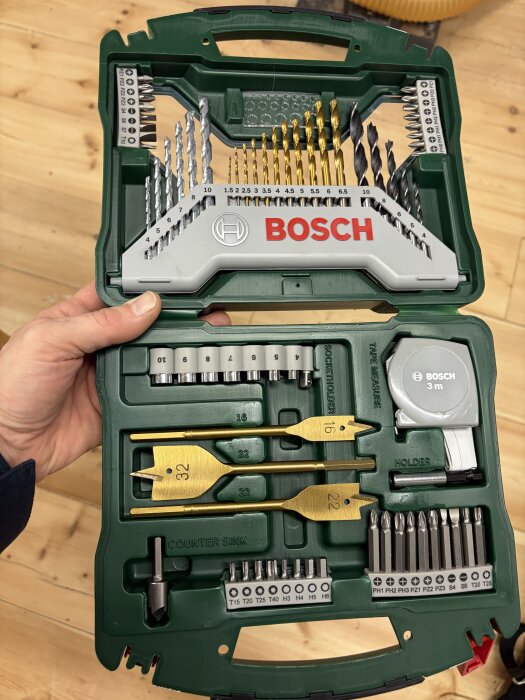 Grön Bosch verktygslåda med olika borrhuvuden, skruvbits och en rullmåttband, håller i hand mot en träbakgrund.