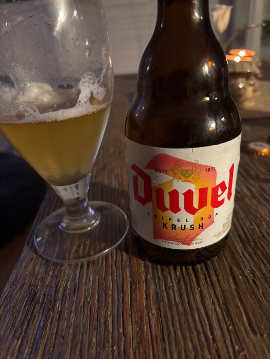Ölflaska av märket "Duvel Tripel Hop Krush" bredvid ett halvtomt glas på ett träbord.
