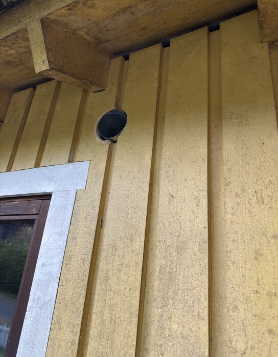 Gult trähus med öppning för ventilationsrör under taket ovanför ett fönster med vit och brun karm.