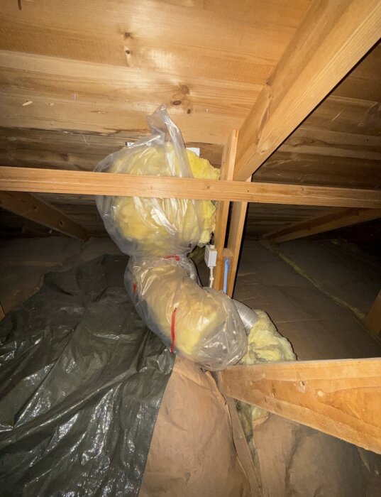Isolerat ventilationsrör på vinden omgivet av plast och tätningsmaterial, del av husets ventilationssystem under renovering.