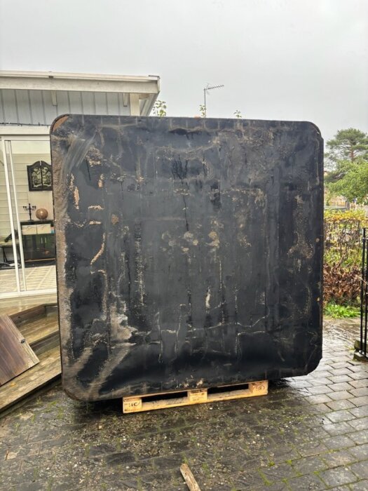 Ett spabad står upprätt på en träpall framför en veranda, efter att ha flyttats för hand med hjälp av rör och en hemmabyggd skateboard.