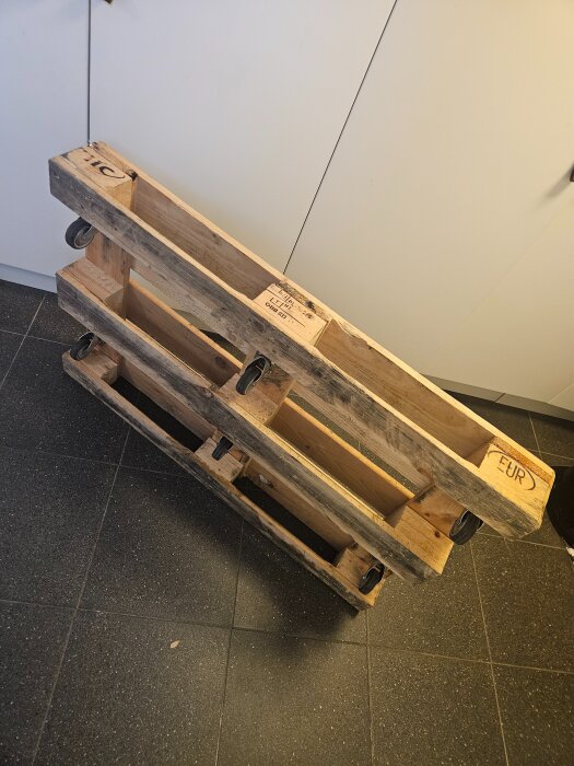 Träpall på hjul, använd som hemmabyggd skateboard för att flytta spabad, står lutad mot en vägg inomhus.