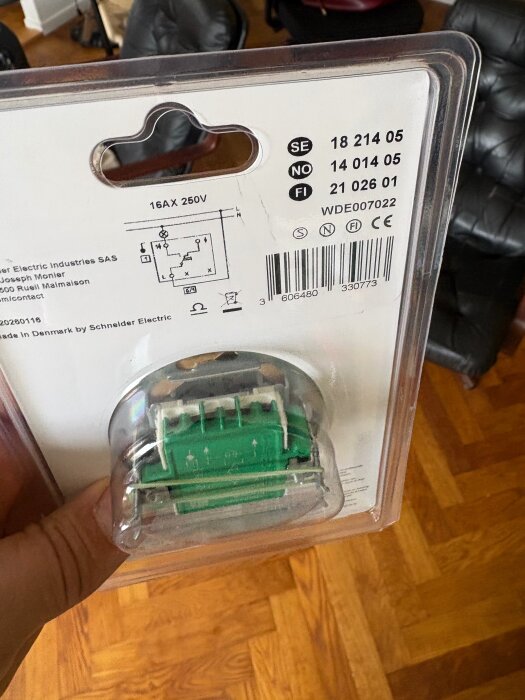 Förpackning med en 16A 250V elektrisk komponent, delnummer SE 18 214 05, från Schneider Electric, visas av en hand på en parkettgolv.