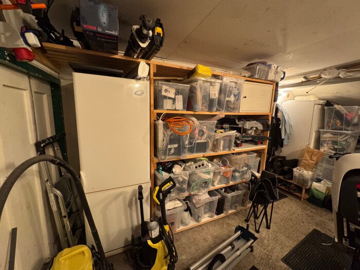 Hylla i garage med plastlådor fyllda med diverse föremål, kyl/frys till vänster; ordning och förvaring i fokus.