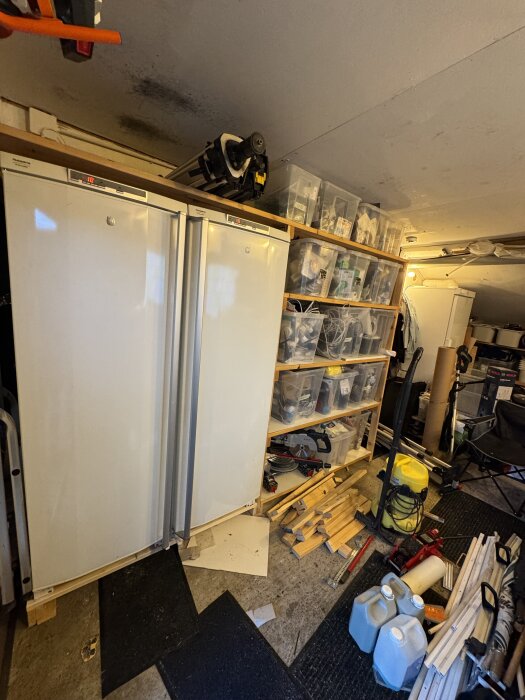 Två vita kylskåp bredvid en hylla med plastlådor i ett garage, omgiven av träplankor och verktyg på golvet.