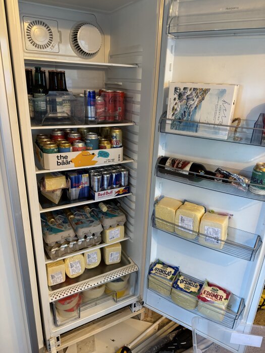 Kylskåp fyllt med drycker, ost och ägg. Hyllorna är organiserade med olika produkter, främst öl, energidrycker och stora block ost.