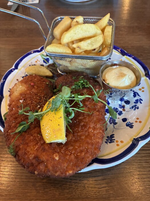 Panerad schnitzel med citron och örter på en tallrik, serverad med pommes frites i metallkorg och en kopp sås.