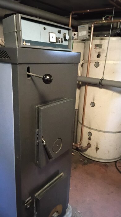 Vedpanna i källare med kopparrör och tank i bakgrunden.