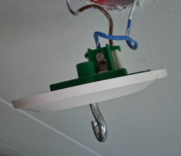 Montering av elektrisk takkontakt med blå och bruna kablar mot ett vitt tak, utrustad med en metallkrok för upphängning.