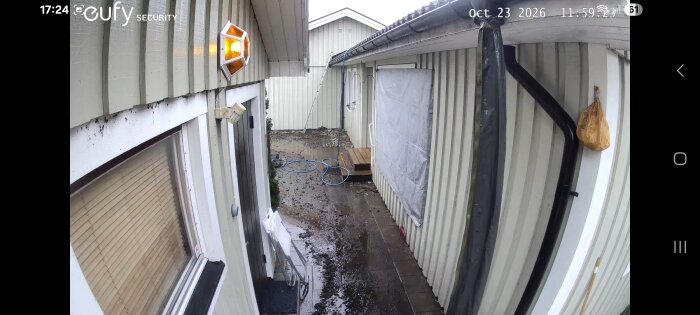 Fasad på hus med täckta fönster och altanöppning, fuktig mark och hängande plastpåse på vägg, utomhusmiljö.