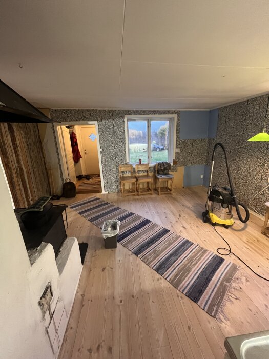 Renoverat kök med trägolv, randig matta, tre stolar vid fönster, vedspis till vänster, dammsugare till höger, och en öppen dörr till ett annat rum.
