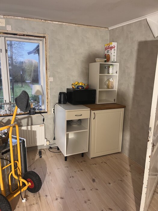 Minikök i vardagsrummet med ett litet skåp, mikrovågsugn, vattenkokare och en kartong flingor ovanpå, med ett fönster till vänster.