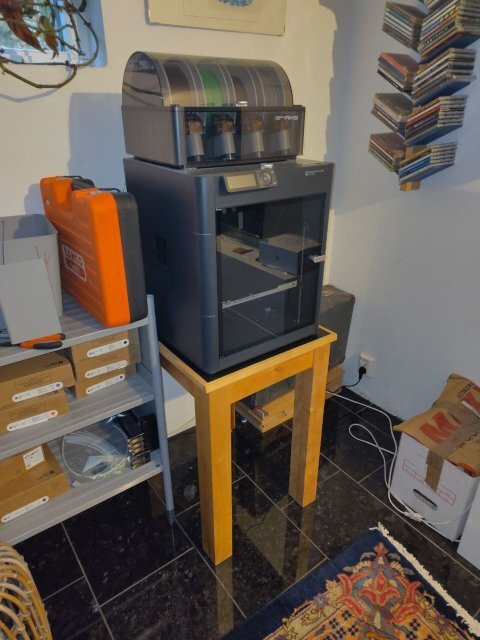 3D-printer placerad på ett hemmabyggt bord i ett rum med hyllor och kartonger.
