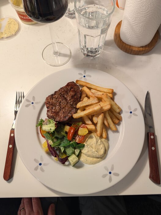Grillad oxfilé med pommes frites, sallad och sås på en tallrik, serverad med glas vin och vatten på ett bord.