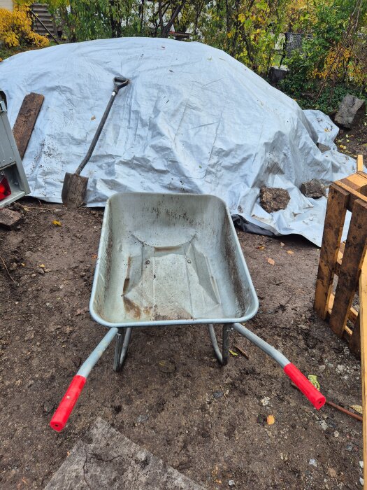 En skottkärra med röda handtag på en jordig byggarbetsplats, bakom den en presenning med en spade lutad mot.
