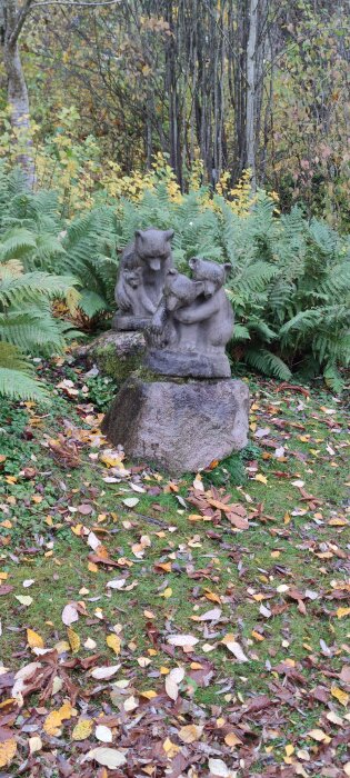 Stenskulptur av två björnar i en skogsmiljö, omgiven av ormbunkar och höstlöv.