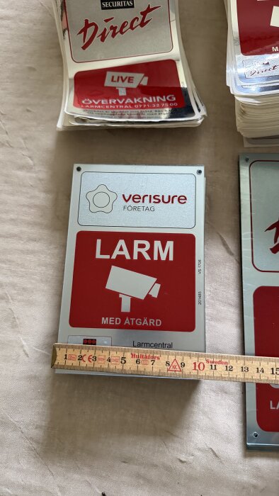 Säkerhetsskyltar för övervakning och larm från Verisure och Securitas Direct, placerade på ett bord med måttstock.