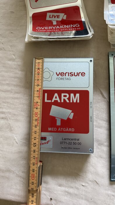 Hårdplast skylt från Verisure Företag med texten "Larm med åtgärd" och telefonnummer, bredvid en linjal som mäter skyltens storlek.
