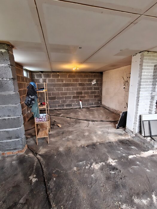Ojämnt betonggolv i en källare med synliga betongväggar, en trähylla och diverse verktyg. Tjära syns på golvet.