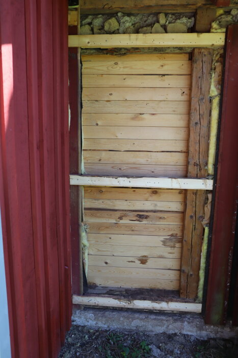 Röd träfasad med lockpanel och blottade, vertikala brädor på baksidan visar vatteninträngning vid kvistar, del av renoveringsprojekt.