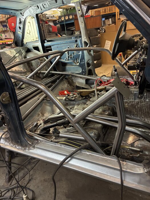 Pågående montering av störtbur i bilens chassi, med verktyg och kablar utspridda i garaget.