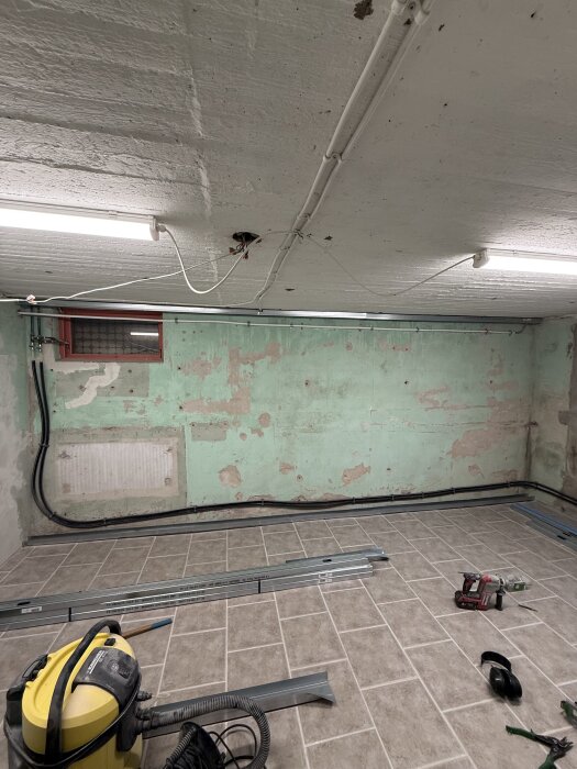Renovering av källarrum med gröna väggar som saknar färg på vissa ställen, tak med synliga elkablar, nya golvplattor, verktyg och dammsugare.