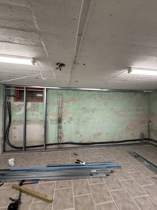 Källare under renovering med synliga reglar på vägg och tak, verktyg och byggmaterial på golvet samt delvis avskalad vägg.