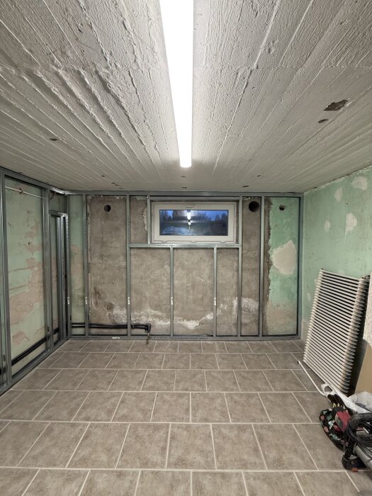 Renovering pågår i ett rum med exponerade väggar, metallstolpar och nylagda golvplattor. Ett litet fönster och lysrör i taket syns.
