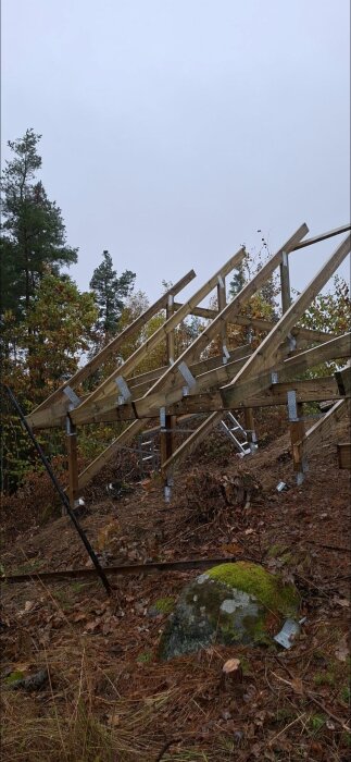 Träram gjord av reglar med metallfästen för en byggstruktur i skogsmiljö. Detaljer som stege och sten synliga i bakgrunden.