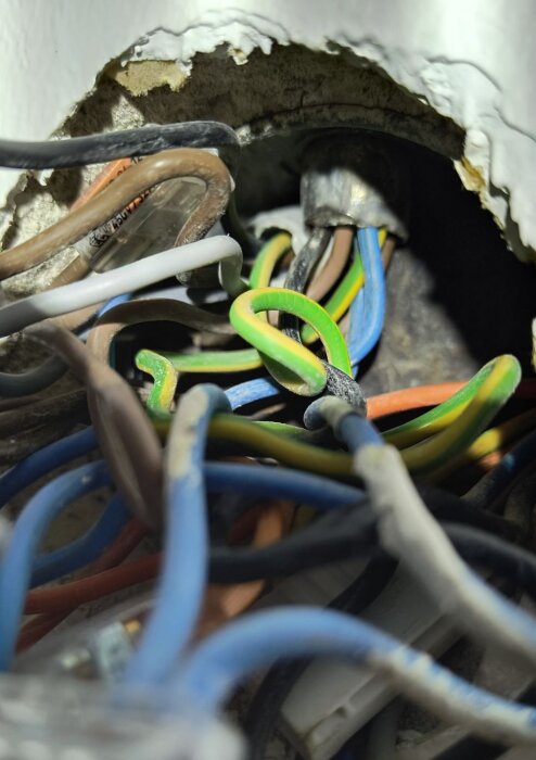 Elektriska ledningar av olika färger trängs i ett vägguttag, inklusive blå, bruna, svarta, grå och gulgröna ledare, i en gammal byggnad.