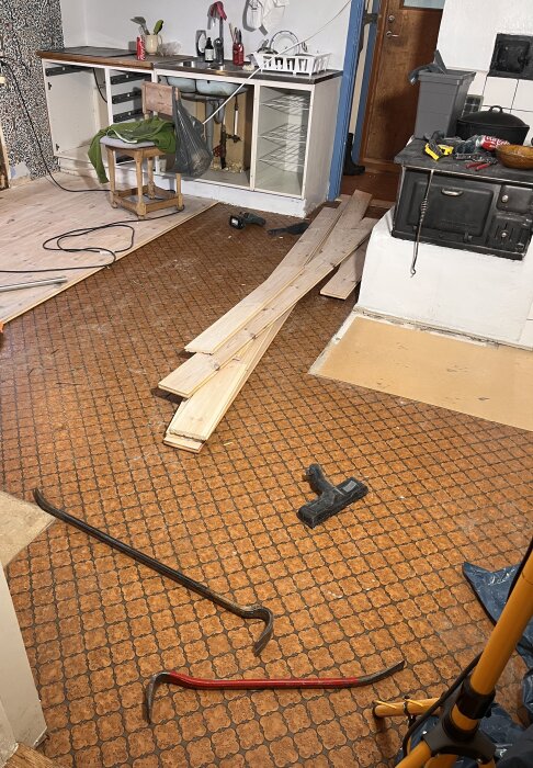 Kök med upptaget golv, plastmatta, brädor, en gammal vedspis och verktyg som kofot och skruvdragare ligger framme under renovering.