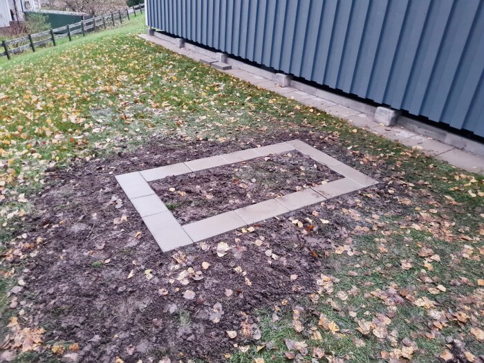Rektangulär ram av markstenar på gräsmatta, omgiven av höstlöv vid en blå byggnad.