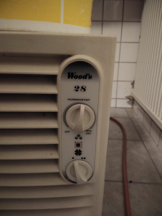 En avfuktare med två reglage för fuktighet och hastighet, märkt "Wood's 28", placerad i en tvättstuga med kakelvägg.