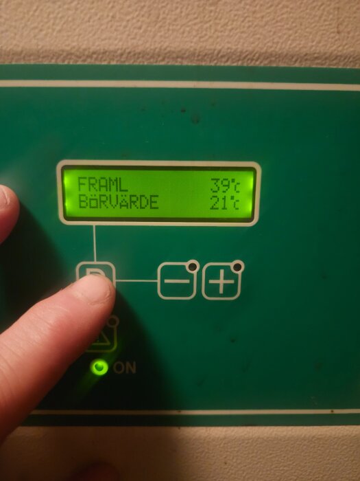 Display med temperaturinställning, "Framl" 39°C och "Börvärde" 21°C, på värmeanläggning. Finger pekar på knapp vid startindikator.