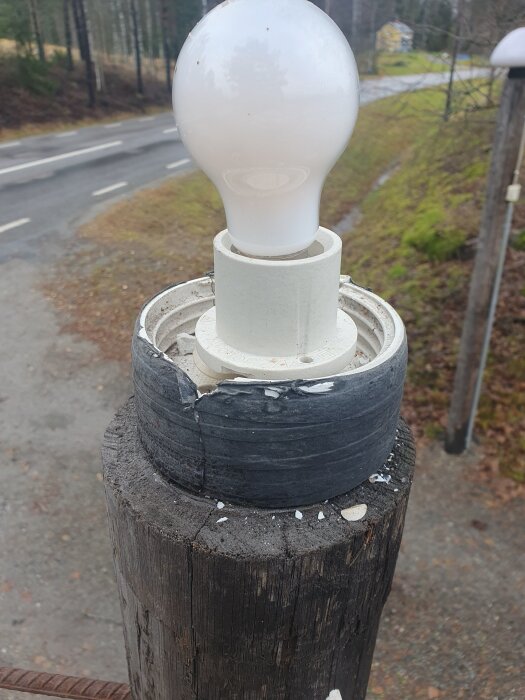 Trasig porslinssockel med svart vulktejp omsluter en glödlampa på en trästolpe vid infart, med närliggande väg i bakgrunden.