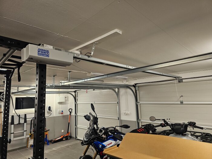 Garageinteriör med garageportsmotor och skena monterade i taket. Motorcyklar parkerade under. Diskussion om att höja motorn för mer utrymme.