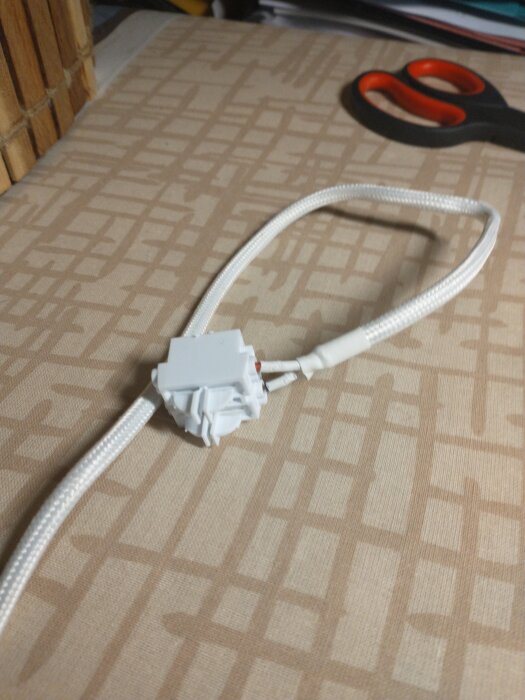 Vit kabel med vävstrumpa ansluten till en vit kopplingsplint (sockerbit) på mönstrad yta, sax i bakgrunden.
