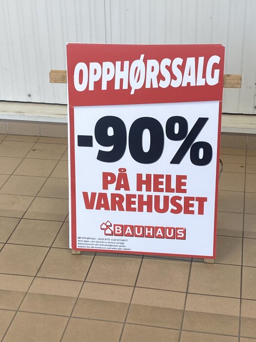 Skylt med texten "OPPHØRSSALG -90% PÅ HELE VAREHUSET" i röd och vit färg från Bauhaus, placerad på ett klinkergolv.