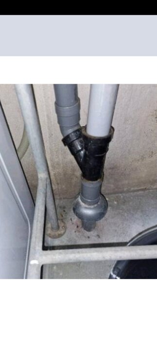 PVC-rör med en Y-koppling och installation mot en betongvägg i ett trångt utrymme, troligen en del av ett avloppssystem i ett hus.