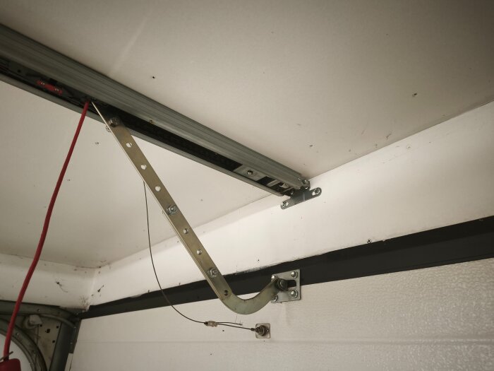 Metallskena och upphängningsmekanism för garageport, sett från insidan med takmonterat spår och röd sladd.