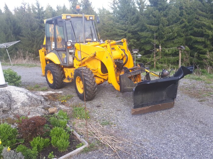 Gul JCB 2cx traktionsgrävare med vikplog på grusväg, omgiven av grönska och en klippa.