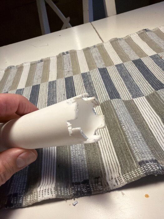Hemgjort verktyg av vitt plastmaterial med skåror, designat för att lossa en ovanlig mutter, hålls i en hand över en randig duk på ett bord.