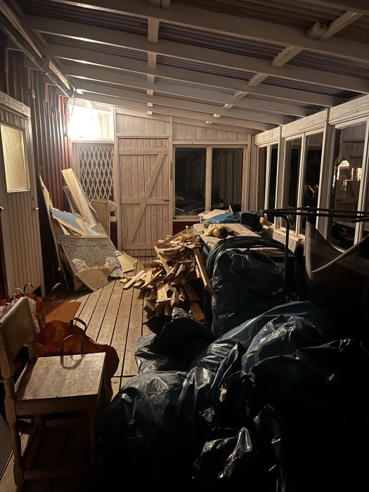 Full veranda med säckar och rivningsmaterial samt staplad bråte från köksrenovering, kvällsbelysning avslöjar träpanel och stora fönster.