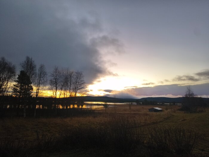 Solnedgång över en flod och äng, med silhuetter av träd i förgrunden och en liten byggnad till höger.