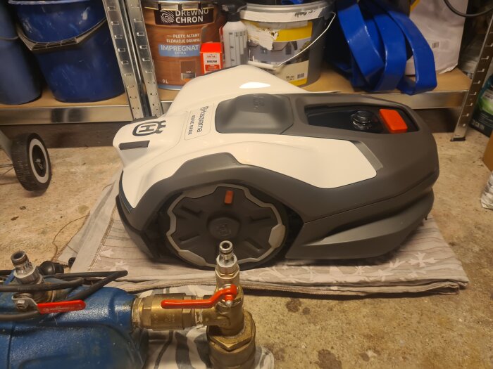 Robotgräsklippare Husqvarna HQ 405ve Nera i garage, omgiven av målburkar och trädgårdsredskap. Ligger på en matta, redo för användning.
