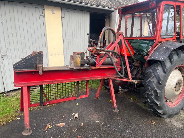 Röd vedklyv kopplad till en traktor står utanför en garagebyggnad.